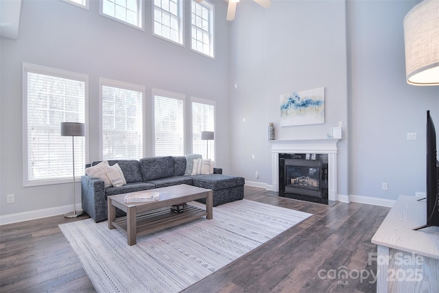 living area with a ceiling fan, dark wood-style floors, a fireplace, baseboards, and a towering ceiling