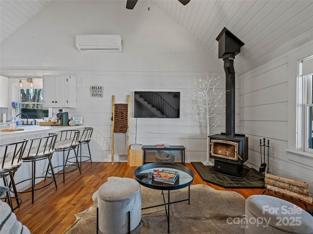 living area with a wall mounted AC, vaulted ceiling, a wood stove, and light wood-type flooring