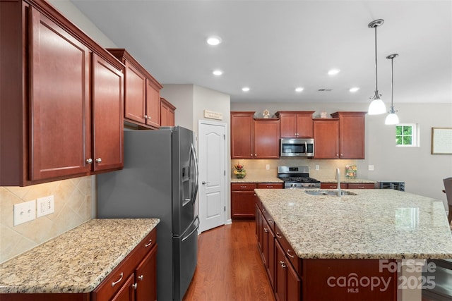 kitchen with reddish brown cabinets, a center island with sink, dark wood finished floors, stainless steel appliances, and a sink