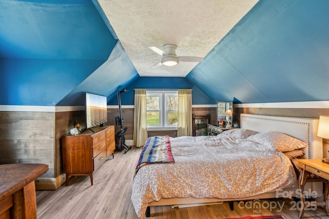 bedroom with light wood-type flooring, a textured ceiling, wood walls, lofted ceiling, and ceiling fan