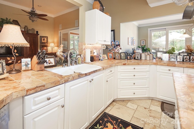 kitchen with stone tile floors, ceiling fan, crown molding, white cabinetry, and a sink