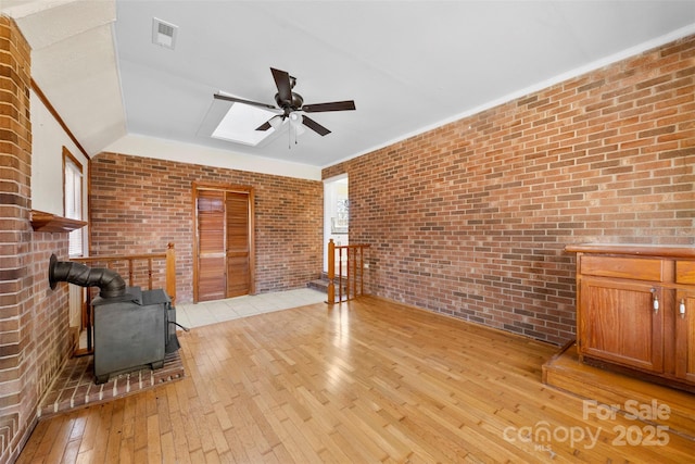 unfurnished living room with light wood finished floors, visible vents, a ceiling fan, a wood stove, and brick wall