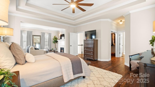 bedroom with dark wood-style floors, baseboards, crown molding, a glass covered fireplace, and a raised ceiling