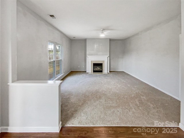 unfurnished living room with ceiling fan, carpet floors, a fireplace with flush hearth, visible vents, and baseboards