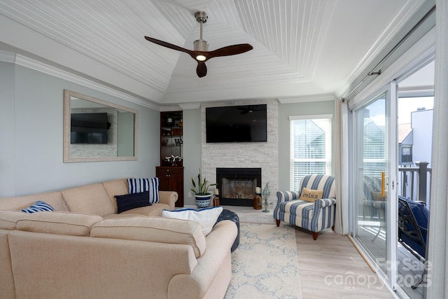 living area with ceiling fan, a stone fireplace, light wood-style flooring, a raised ceiling, and crown molding