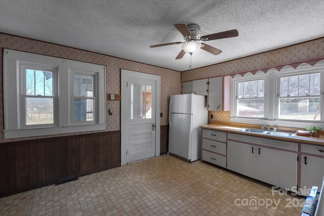 kitchen with freestanding refrigerator, wainscoting, a sink, a textured ceiling, and wallpapered walls