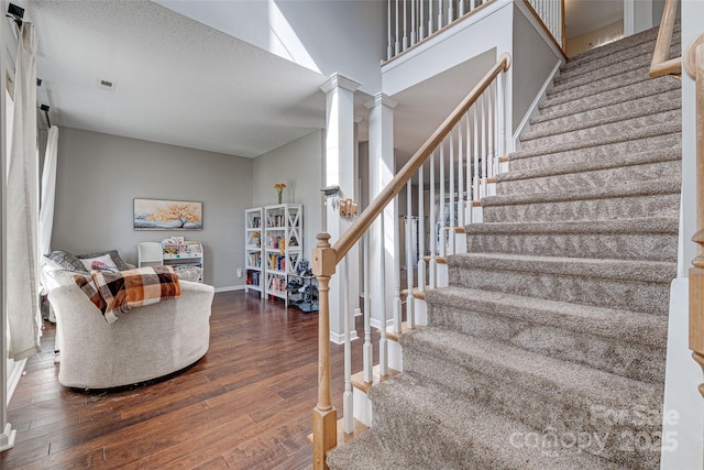 staircase featuring baseboards, visible vents, wood-type flooring, a high ceiling, and ornate columns