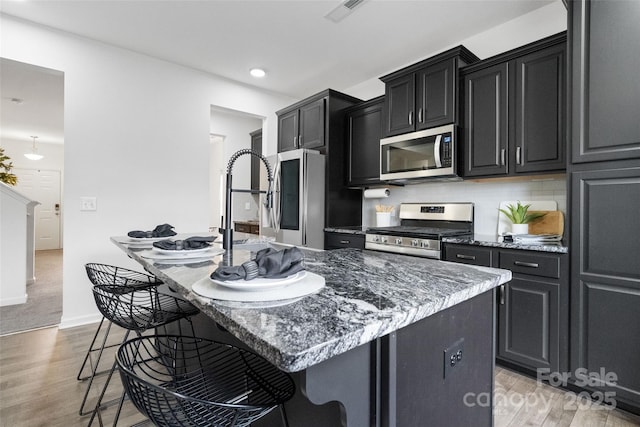 kitchen with a breakfast bar area, visible vents, appliances with stainless steel finishes, a kitchen island with sink, and dark cabinets