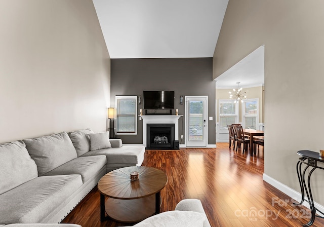 living area with baseboards, a fireplace with flush hearth, wood finished floors, a notable chandelier, and high vaulted ceiling