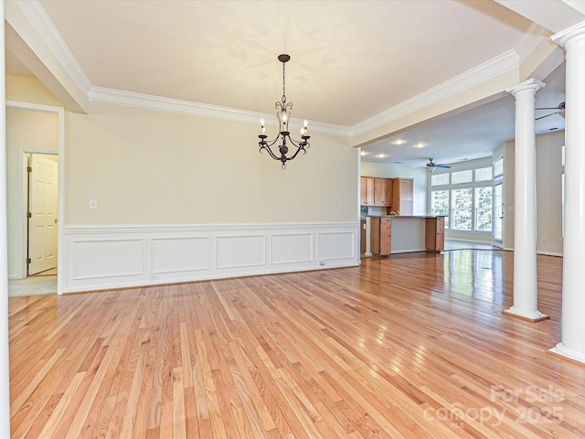 empty room featuring a wainscoted wall, ornamental molding, decorative columns, ceiling fan with notable chandelier, and light wood-style flooring