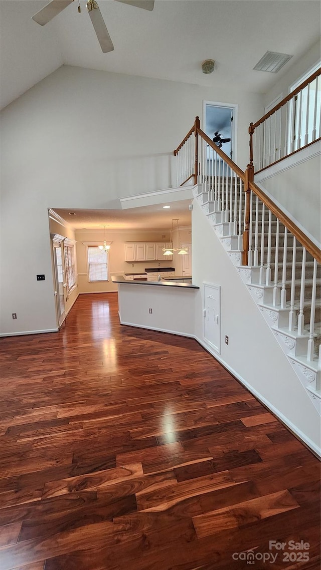unfurnished living room with high vaulted ceiling, ceiling fan with notable chandelier, dark wood-style flooring, visible vents, and stairway