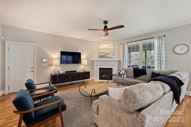 living area featuring light wood-style floors, a glass covered fireplace, ceiling fan, and baseboards