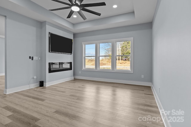 unfurnished living room featuring baseboards, a raised ceiling, a glass covered fireplace, light wood-style flooring, and recessed lighting