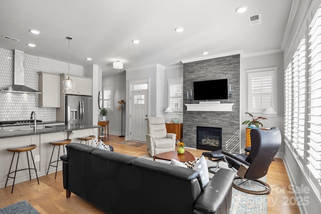 living room with light wood-type flooring, visible vents, a stone fireplace, and crown molding