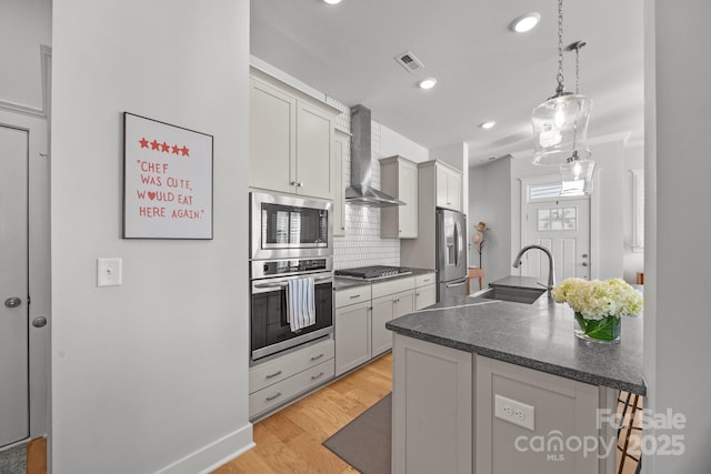 kitchen featuring light wood finished floors, dark countertops, wall chimney range hood, appliances with stainless steel finishes, and a sink