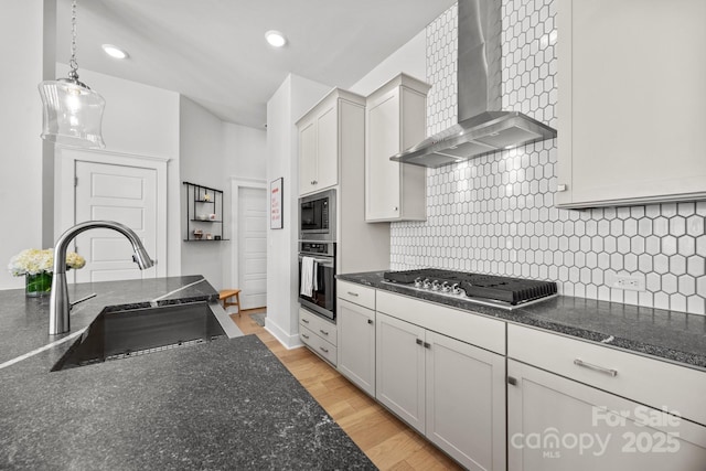 kitchen with a sink, decorative light fixtures, wall chimney range hood, appliances with stainless steel finishes, and light wood finished floors