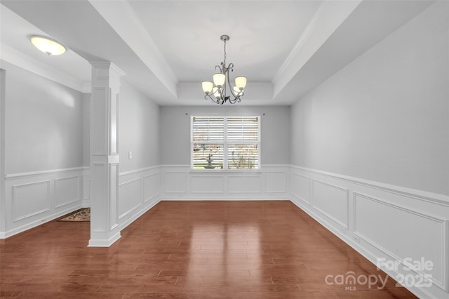 unfurnished dining area with decorative columns, a raised ceiling, wood finished floors, crown molding, and a chandelier