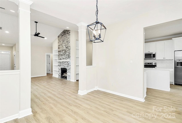 unfurnished dining area featuring light wood-type flooring, ceiling fan with notable chandelier, a large fireplace, baseboards, and ornate columns