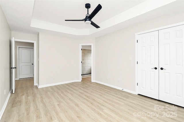 unfurnished bedroom featuring baseboards, ceiling fan, a tray ceiling, light wood-style flooring, and a closet