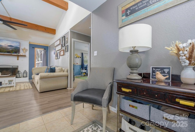 living area featuring ceiling fan, a textured wall, tile patterned flooring, a brick fireplace, and beamed ceiling