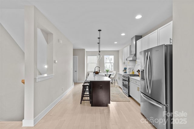 kitchen featuring wall chimney range hood, an island with sink, stainless steel appliances, white cabinets, and a kitchen breakfast bar