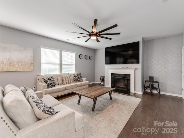 living area with dark wood-style flooring, a fireplace with flush hearth, a ceiling fan, baseboards, and wallpapered walls