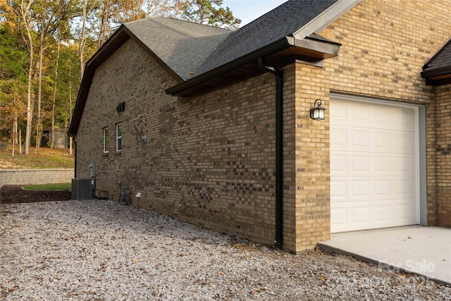 view of side of property featuring a garage, central air condition unit, a shingled roof, and brick siding