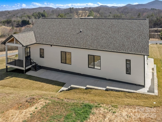 rear view of house featuring a mountain view, roof with shingles, a patio, and a lawn