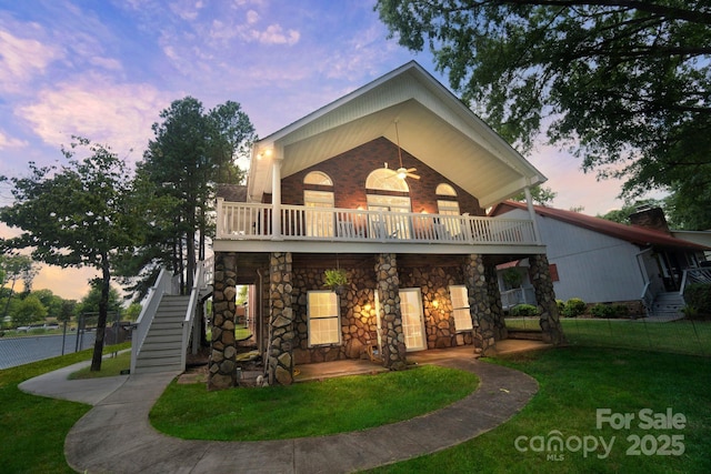 back of property at dusk with ceiling fan, a balcony, brick siding, stairs, and a yard