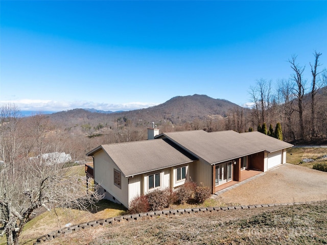 view of front of home with dirt driveway, a view of trees, a chimney, an attached garage, and a mountain view