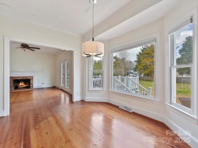 unfurnished dining area featuring a warm lit fireplace, a ceiling fan, visible vents, baseboards, and hardwood / wood-style floors