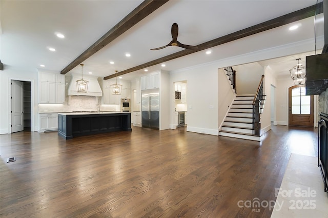 unfurnished living room featuring baseboards, dark wood-type flooring, stairs, beam ceiling, and ceiling fan with notable chandelier