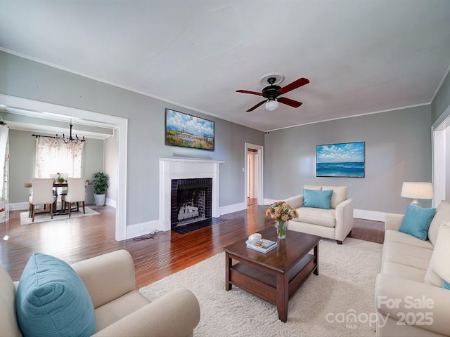 living room featuring baseboards, ornamental molding, wood finished floors, a brick fireplace, and ceiling fan with notable chandelier