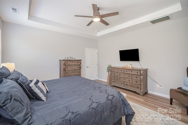 bedroom with visible vents, baseboards, light wood-type flooring, a tray ceiling, and a ceiling fan