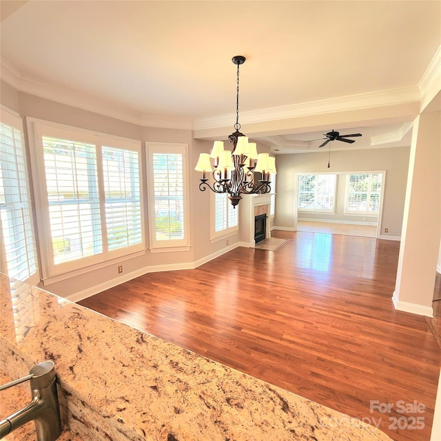 unfurnished dining area with ceiling fan with notable chandelier, crown molding, a fireplace with flush hearth, and wood finished floors