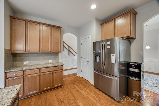 kitchen featuring stainless steel refrigerator with ice dispenser, arched walkways, light wood finished floors, decorative backsplash, and light stone countertops