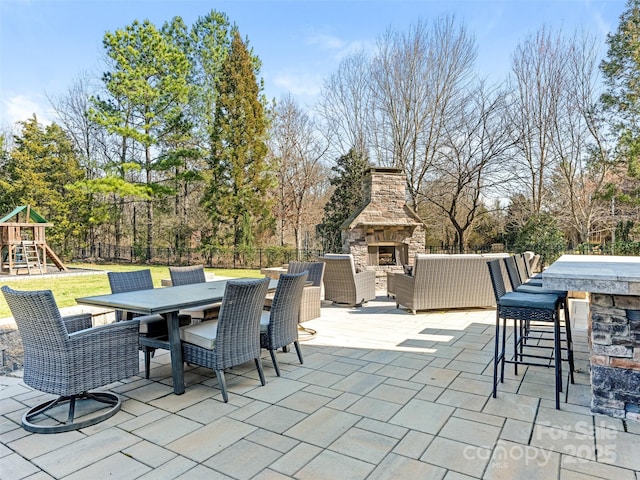 view of patio / terrace with outdoor dry bar, an outdoor living space with a fireplace, a playground, fence, and outdoor dining area