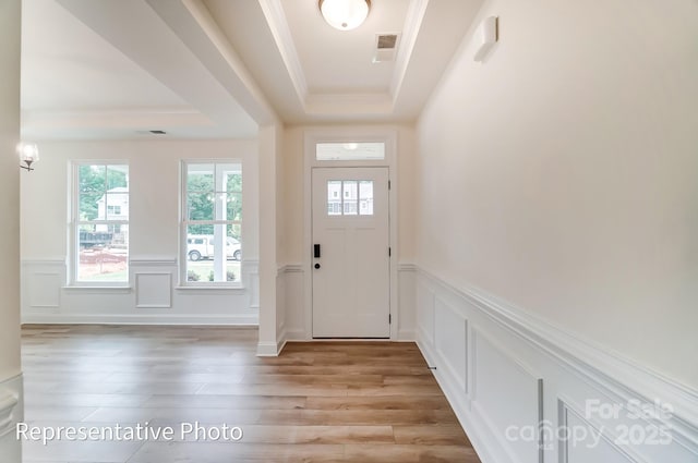 foyer with light wood-type flooring, visible vents, a tray ceiling, wainscoting, and crown molding