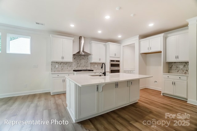 kitchen featuring visible vents, a kitchen island with sink, a sink, white cabinets, and wall chimney range hood