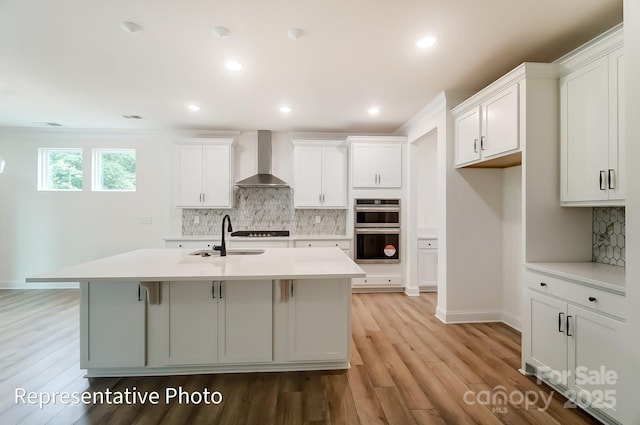 kitchen with a sink, wall chimney range hood, backsplash, double oven, and light countertops