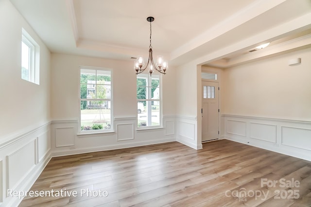 unfurnished dining area featuring a notable chandelier, wainscoting, light wood-type flooring, and a raised ceiling