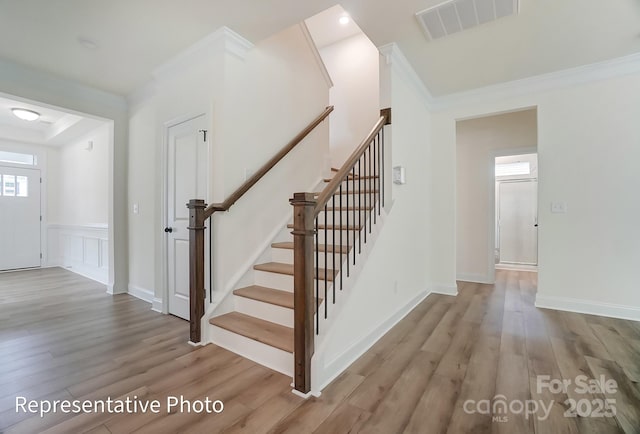 stairs with wood finished floors, a wainscoted wall, baseboards, visible vents, and ornamental molding