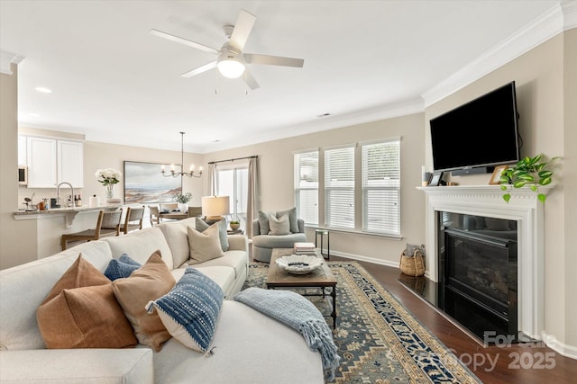 living area featuring baseboards, a fireplace with flush hearth, ornamental molding, dark wood-type flooring, and ceiling fan with notable chandelier