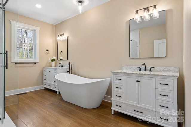 full bath featuring baseboards, two vanities, wood finished floors, a soaking tub, and a sink