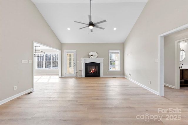 unfurnished living room with high vaulted ceiling, light wood-style floors, a warm lit fireplace, and ceiling fan