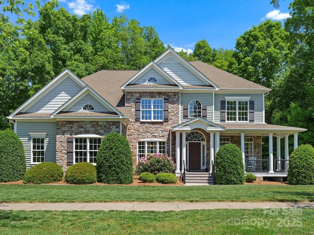view of front facade featuring a shingled roof, a front yard, stone siding, and covered porch