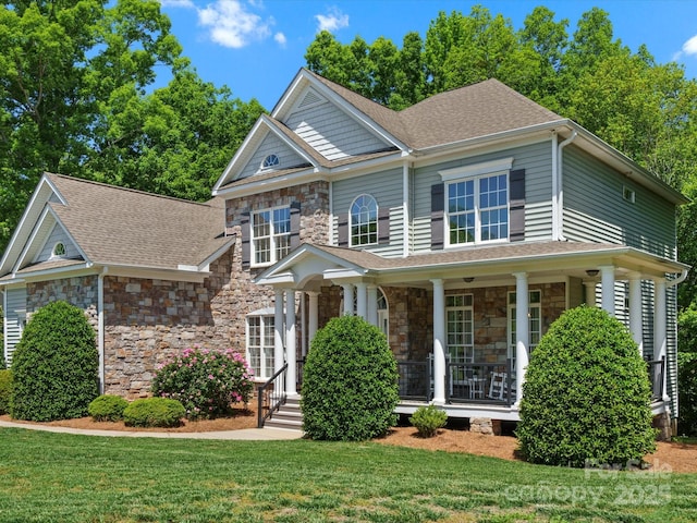 view of front facade with a porch, roof with shingles, a front yard, and stone siding