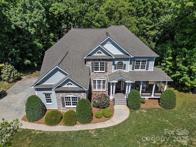 view of front of property with covered porch, driveway, roof with shingles, a front yard, and stone siding