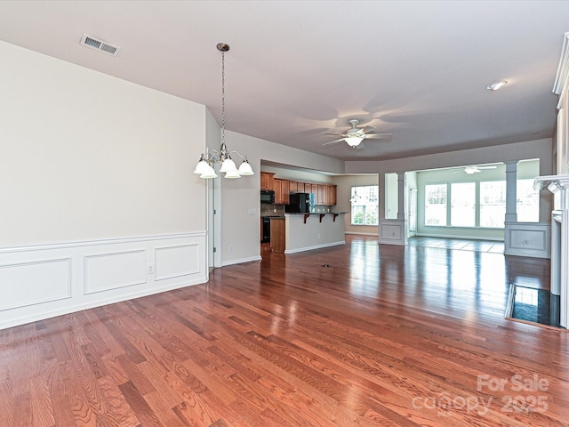 unfurnished living room with wood finished floors, a wainscoted wall, visible vents, ornate columns, and ceiling fan with notable chandelier