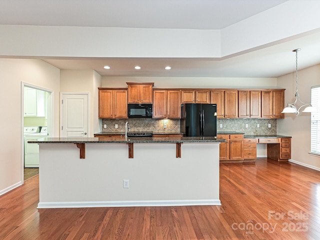 kitchen featuring dark wood finished floors, washer / dryer, brown cabinets, black appliances, and a kitchen island with sink